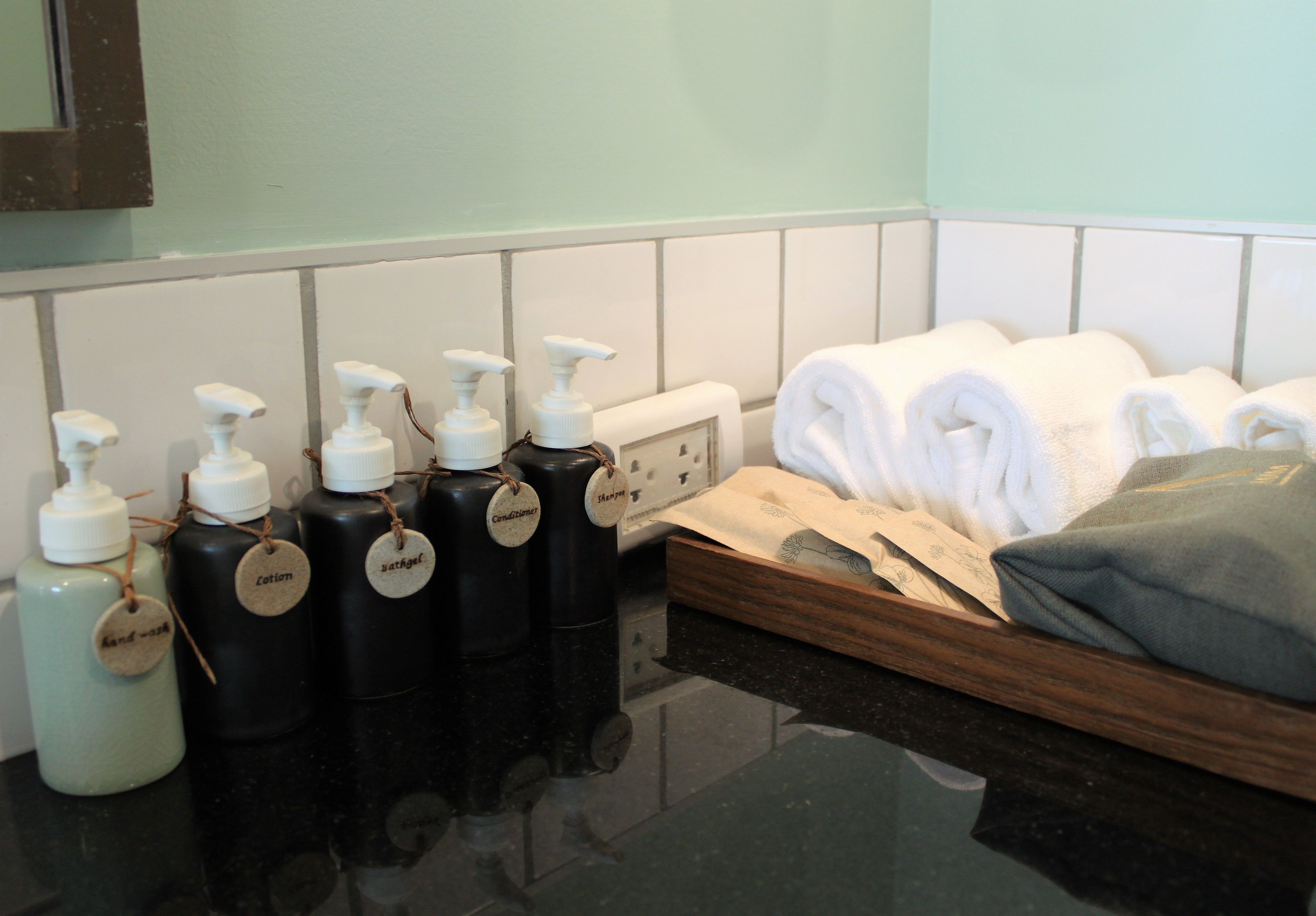 A bathroom with black and white bottles labeled with various amenities, a wooden tray with towels, and a mirror on the wall.