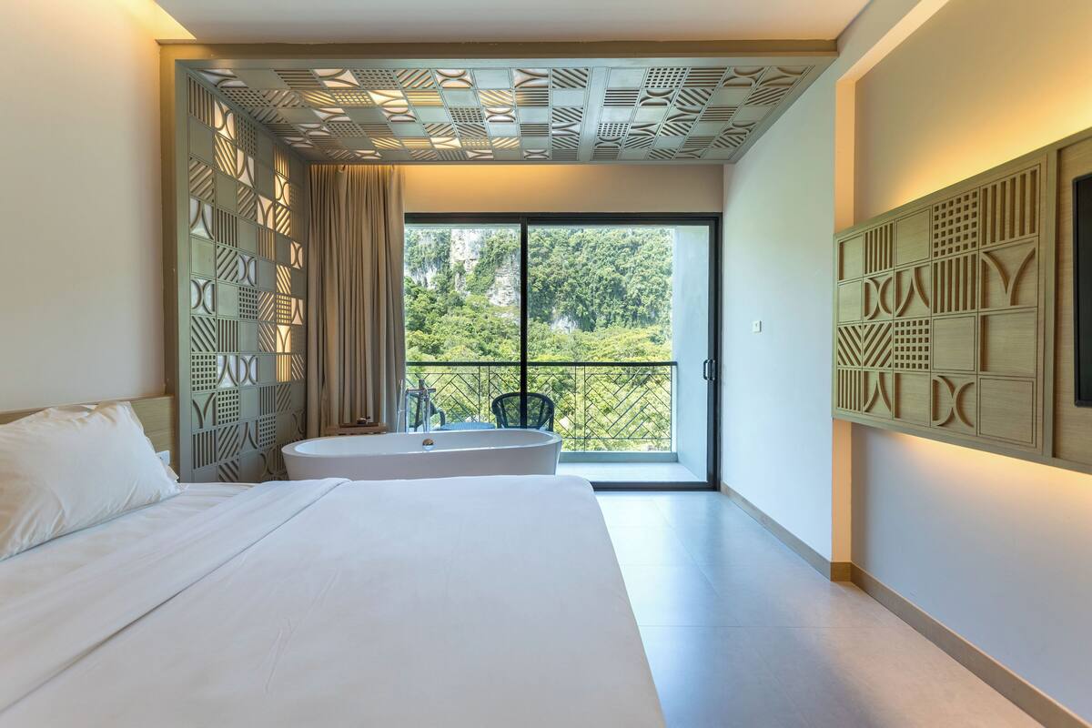 A modern bedroom with a large bed, a built-in bathtub, and a balcony with a view of greenery.