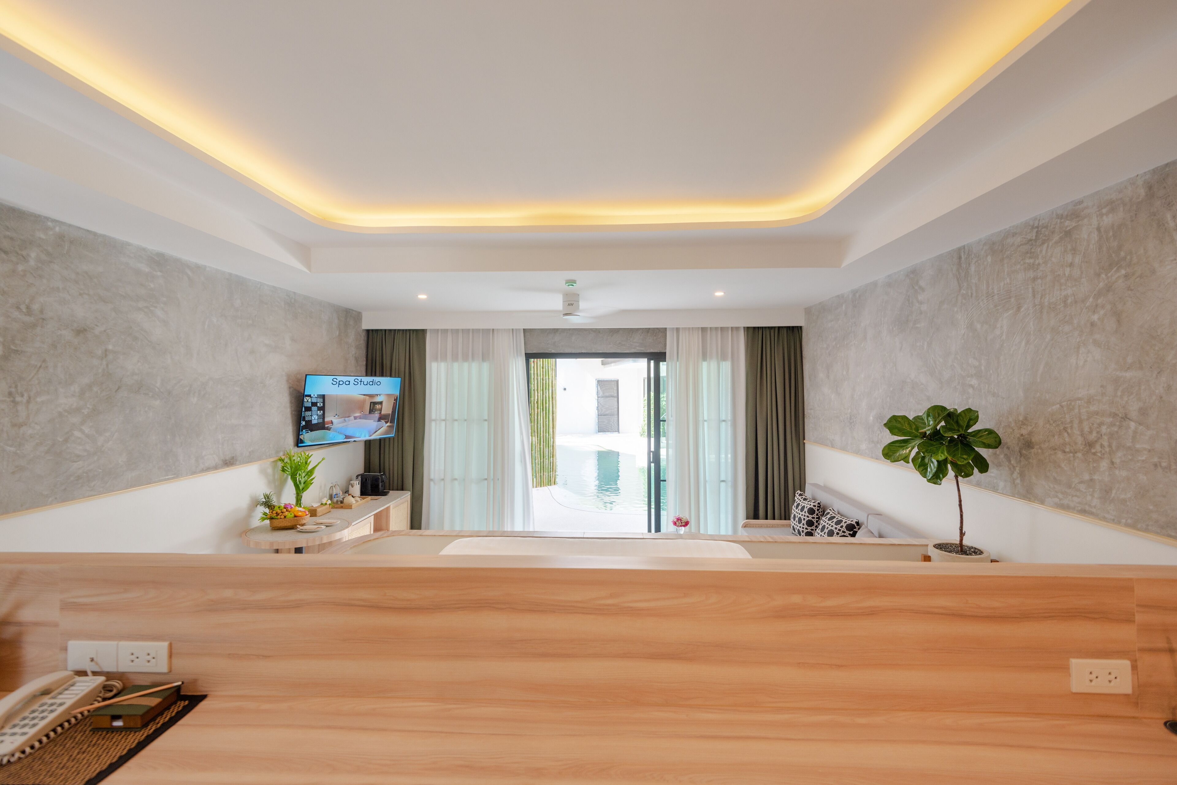 Zona de recepción de un hotel con un mostrador de madera, un televisor mostrando 'Spa Studio' y vista a una piscina a través de puertas de vidrio.