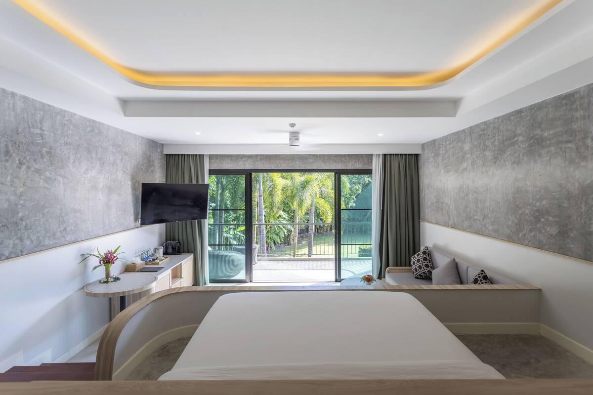 A modern hotel room with a large bed, a flat-screen TV, a small table with a vase of flowers, and a balcony with a view of greenery.