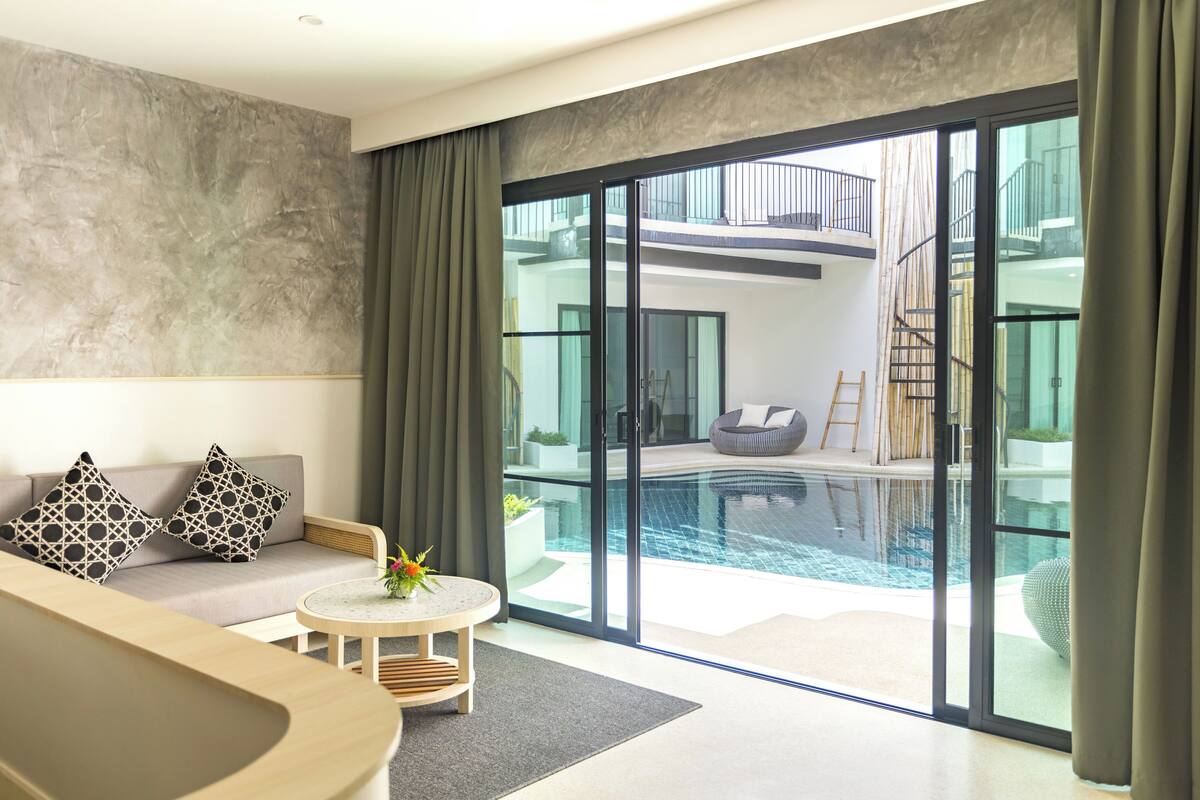 A modern hotel room with a large sliding glass door leading to a pool area, a sofa with patterned cushions, and a small round table with a flower vase.