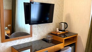 A hotel room with a wooden desk, a television mounted on the wall, a mirror, a small table with a kettle, and a grey chair. - Pinar Elite Hotel (Adana)
