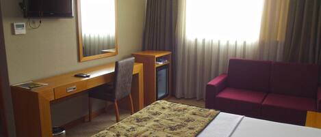A hotel room with a bed, a desk with a chair, a television, a mirror, and a sofa.