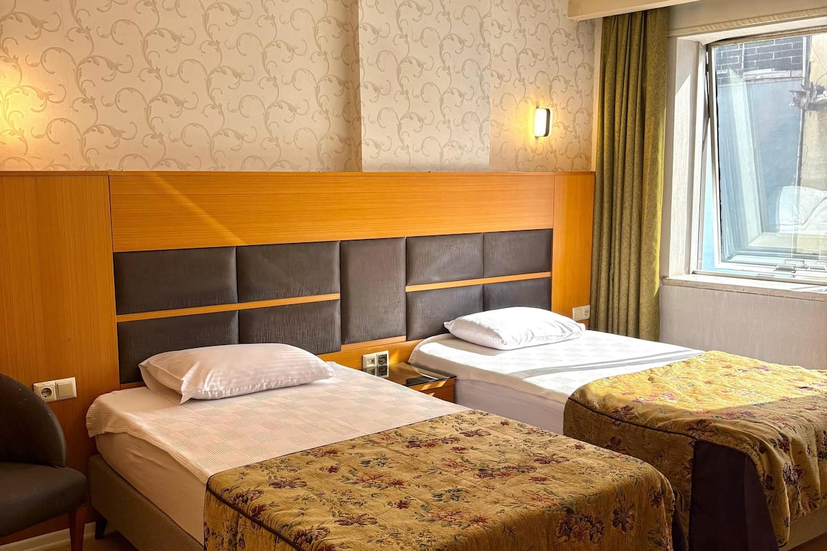A hotel room with two beds, a wooden headboard, a nightstand, a chair, and a window with curtains.