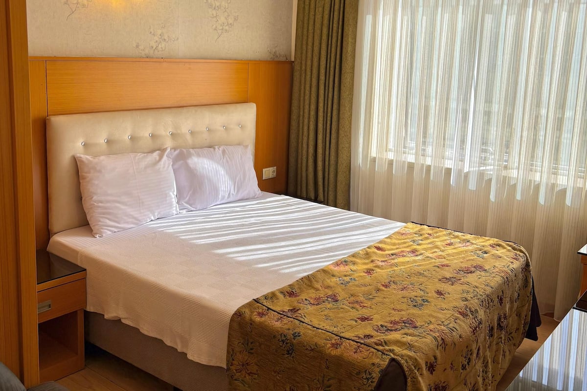 A hotel room with a bed, a nightstand, a chair, a desk, a window with curtains, and a wall-mounted light.
