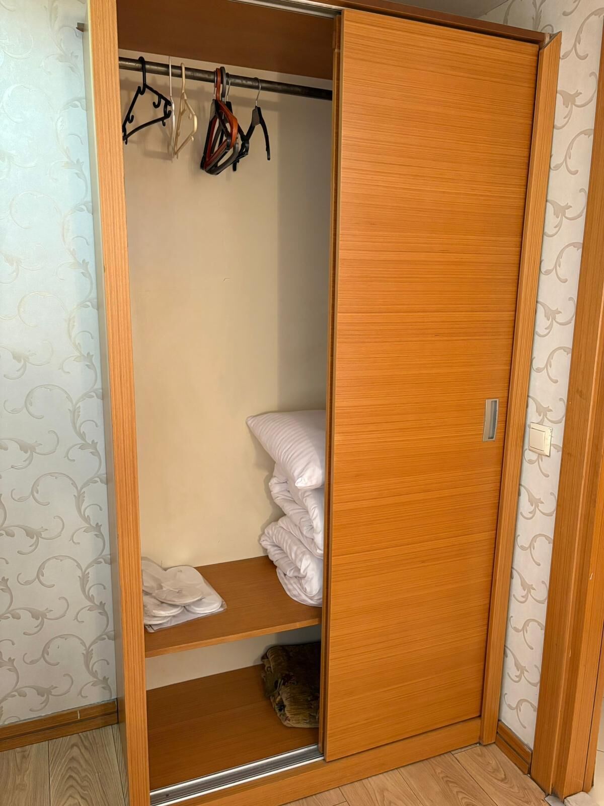 A wooden wardrobe with a sliding door, a hanging rack with clothes, and a bed with white bedding.