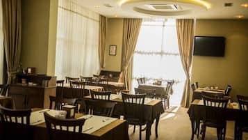 Une salle à manger d’hôtel avec plusieurs tables et chaises, un téléviseur à écran plat fixé au mur et de grandes fenêtres dotées de rideaux.