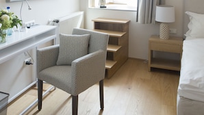A hotel room with a wooden floor, a desk with a lamp, a chair, a bed, and a window with curtains. - Cottage Rheingau Hotel (Walluf)