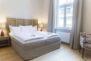 A neatly made bed with white linens and pillows, a wooden headboard, two bedside lamps, a window with curtains, and a patterned armchair. - Cottage Rheingau Hotel (Walluf)
