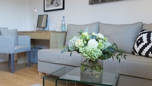A living room with a glass coffee table, a grey sofa, a framed picture on the wall, and a small desk with a chair. - Cottage Rheingau Hotel (Walluf)