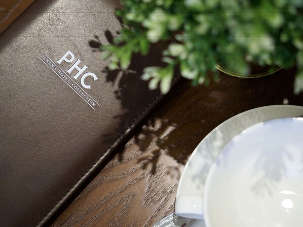 A brown leather-bound book with "PHC" and "PENANG HOTEL COLLECTION" embossed on it, placed on a wooden surface next to a white plate and a glass. - Ropewalk Piazza Hotel Managed By The Ascott Limited (George Town)
