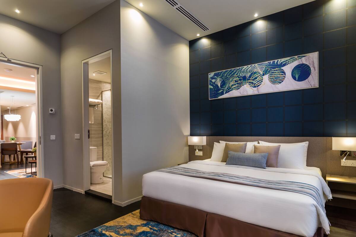 A modern hotel room with a large bed, a bathroom, and a dining area visible through an open door.