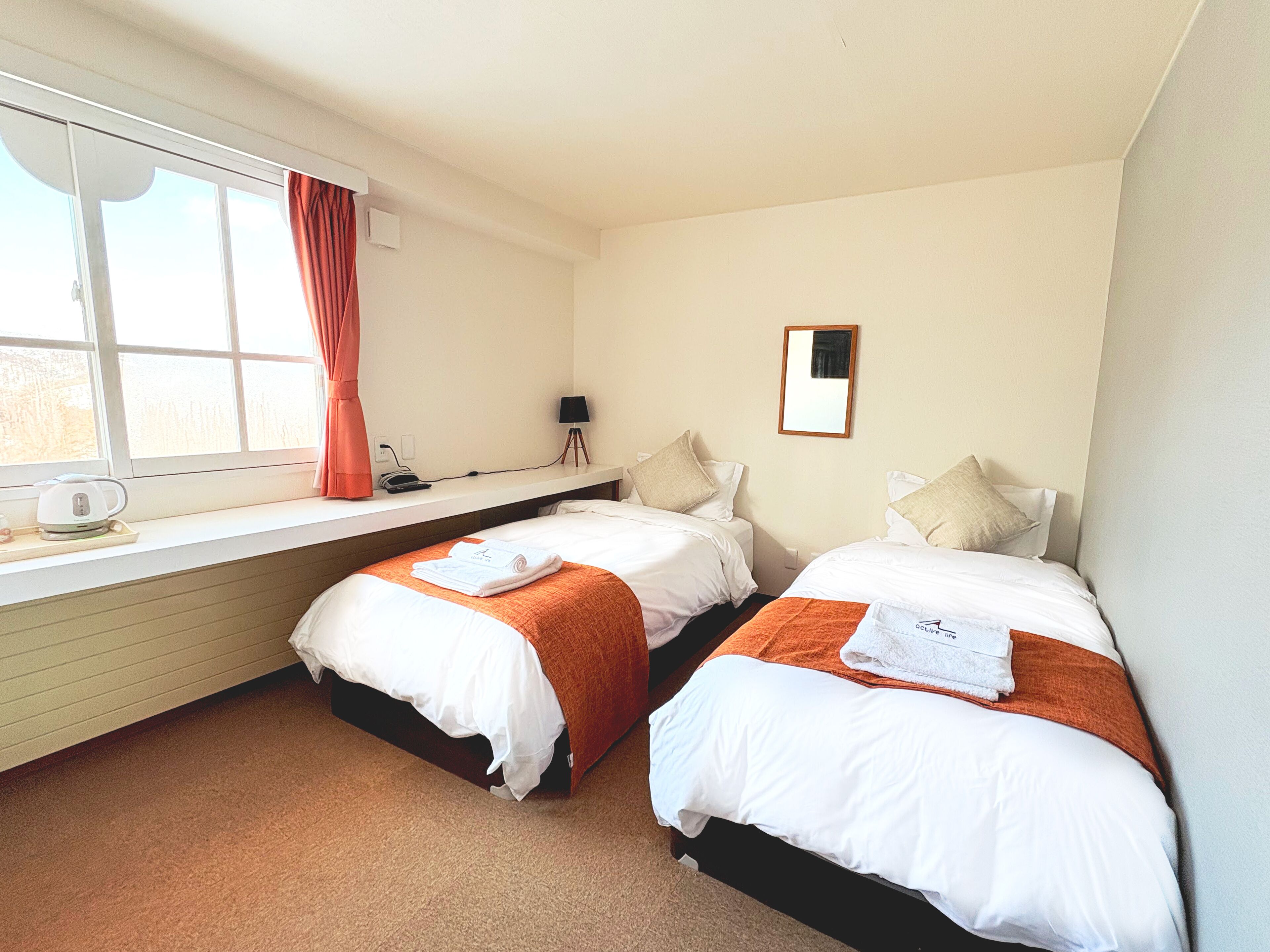 A hotel room with two beds, a window with curtains, a bedside table with a lamp, and a small desk with a kettle.