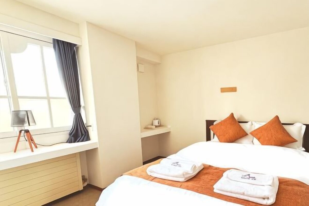 A hotel room with a bed, pillows, a window with curtains, a bedside table, and a lamp.