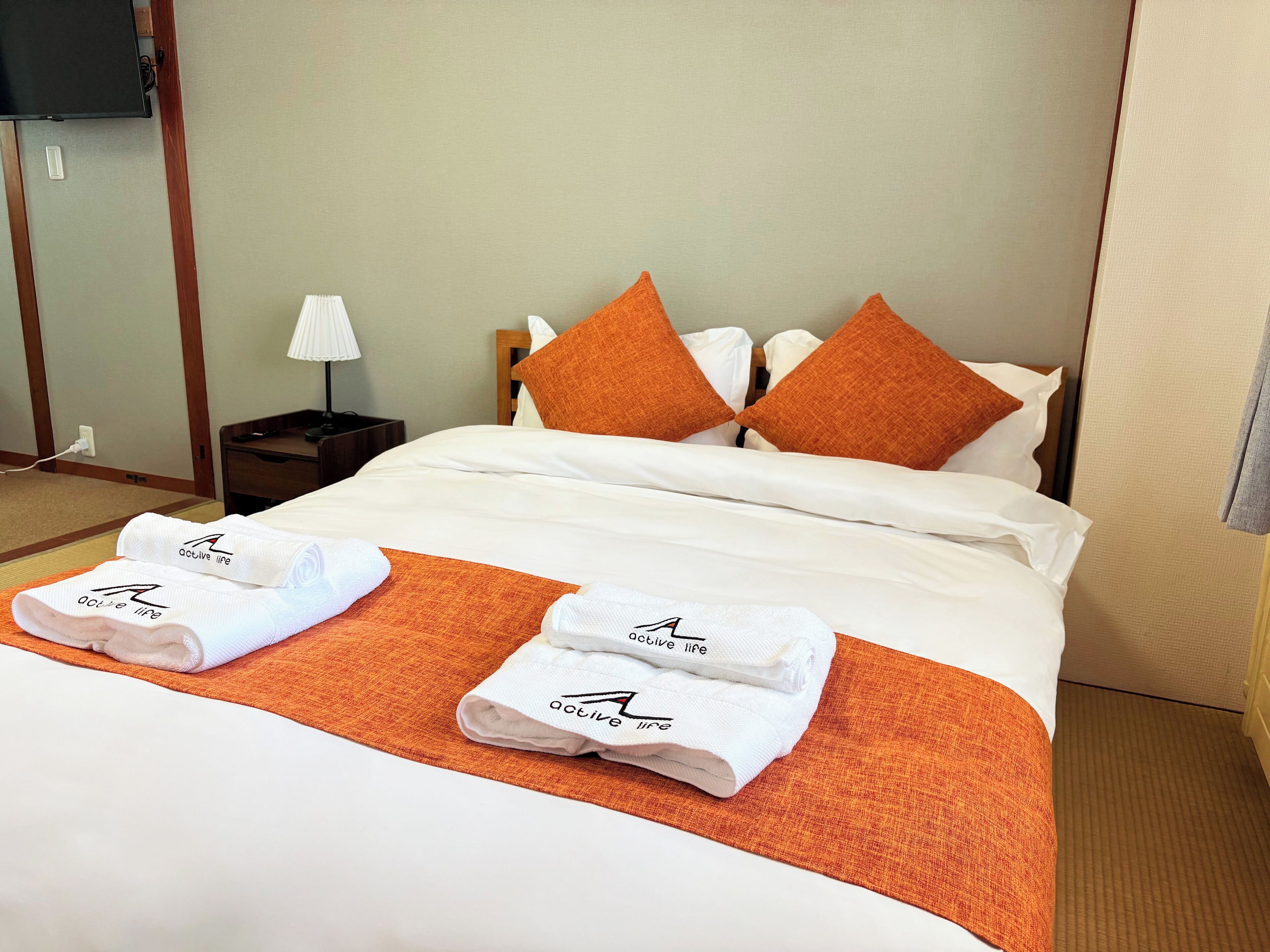 A hotel room with a neatly made bed, a nightstand with a lamp, and a folded towel with a logo on it.