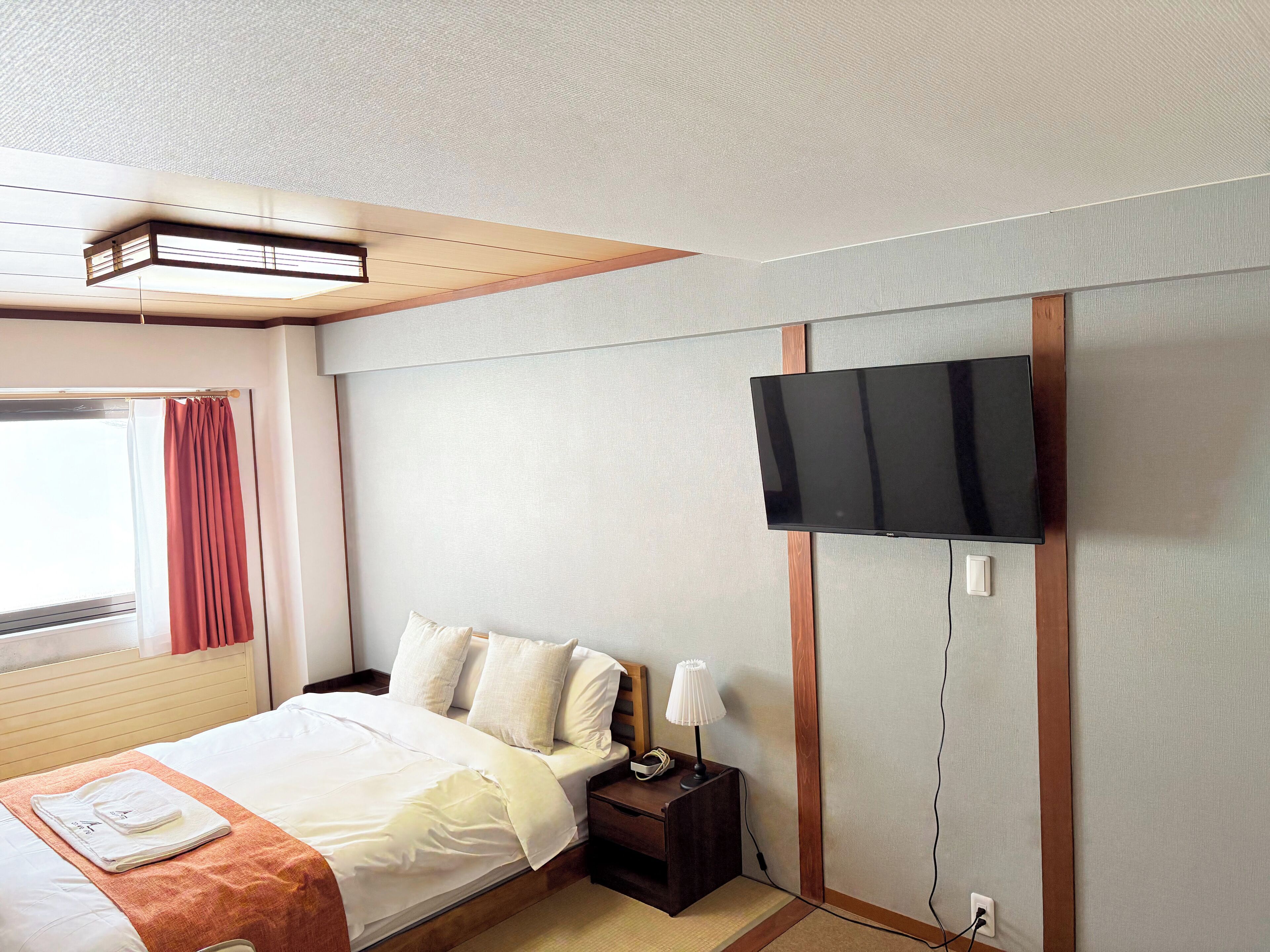 A hotel room with a bed, a bedside table with a lamp, a TV mounted on the wall, and a window with curtains.