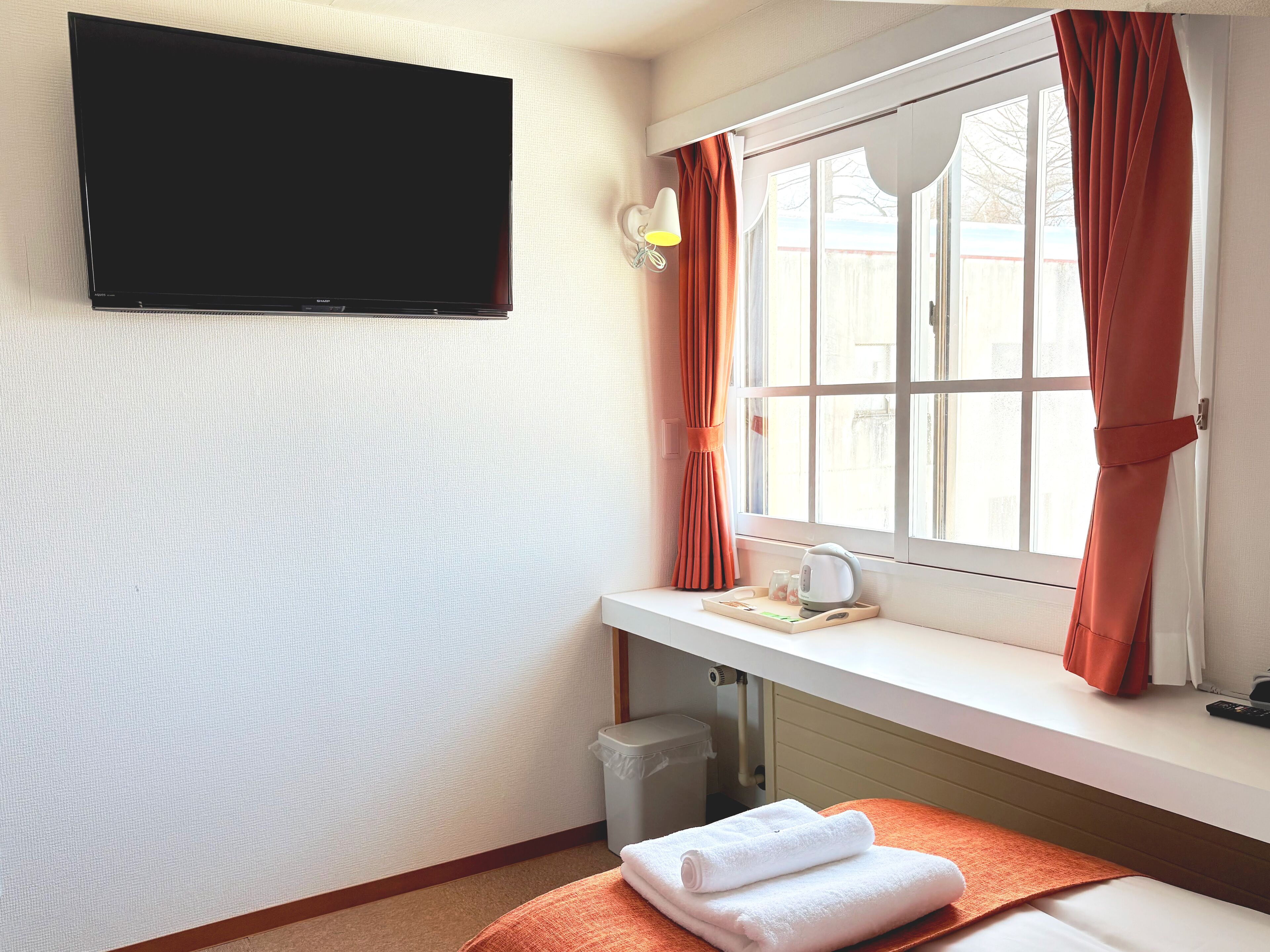 A hotel room with a flat-screen TV mounted on the wall, a window with orange curtains, a bedside table with a kettle and tea set, and a neatly folded white towel on the bed.
