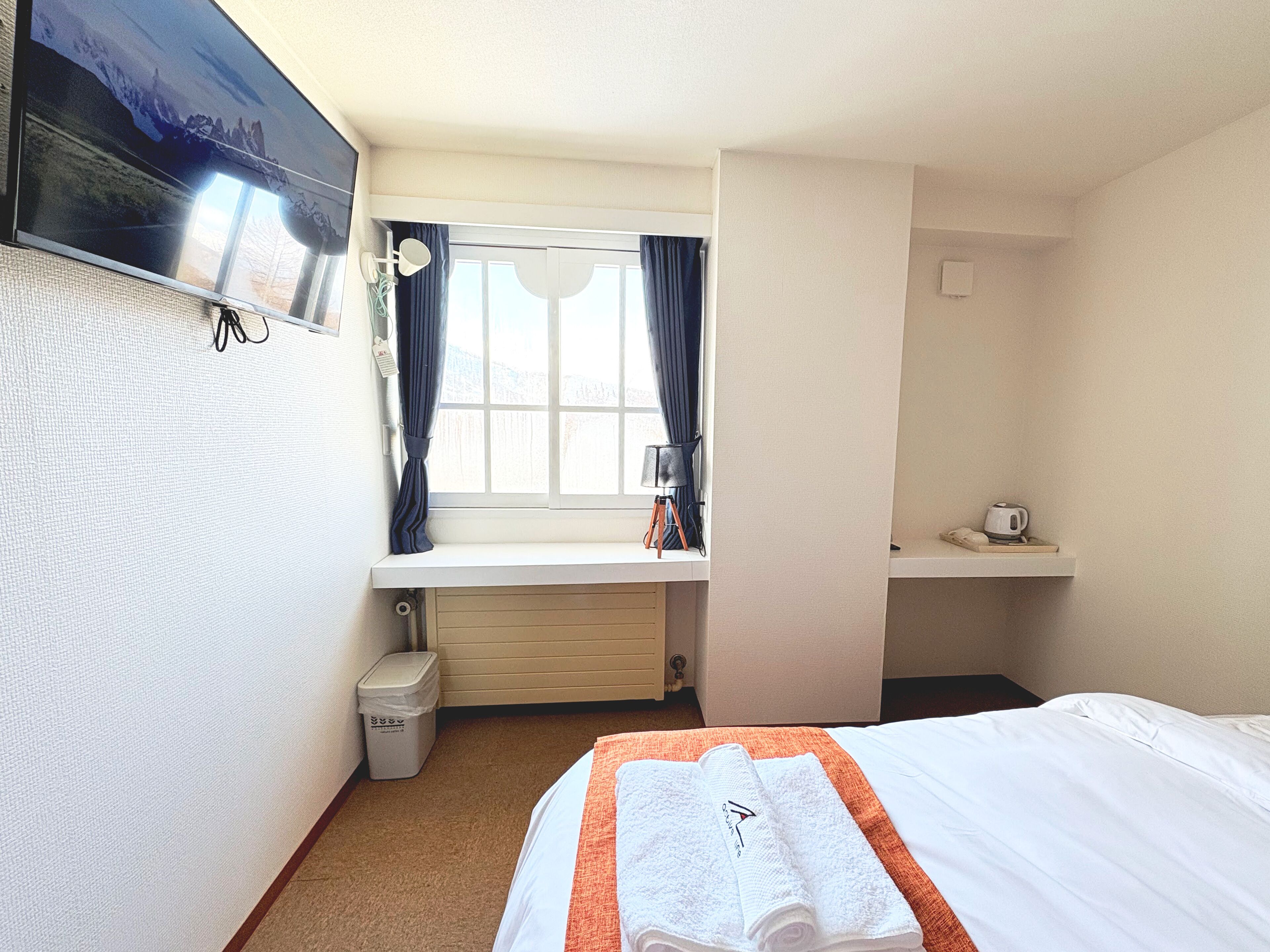 A hotel room with a bed, a window with curtains, a television mounted on the wall, a small table with a kettle, and a trash bin.