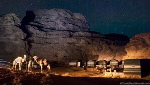 Cielo estrellado con una gran formación rocosa y varias tiendas de campaña o caravanas al frente.