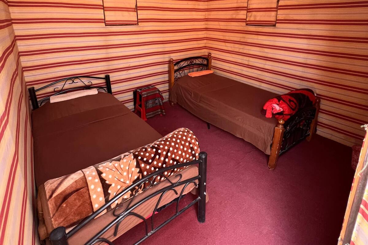 Une petite pièce aux murs rayés, avec deux lits, une petite table et un tapis rouge.