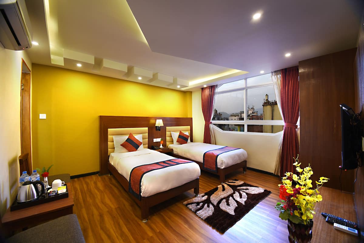 A hotel room with two beds, a wooden floor, a large window with a city view, a small table with a tray of items, and a vase of flowers.