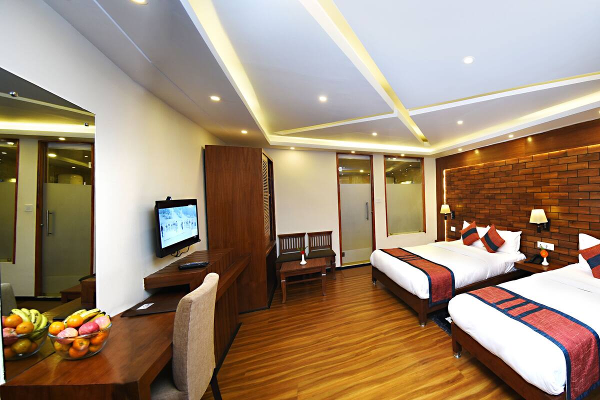 A hotel room with two beds, a desk with a TV, a fruit bowl, and a wooden floor.