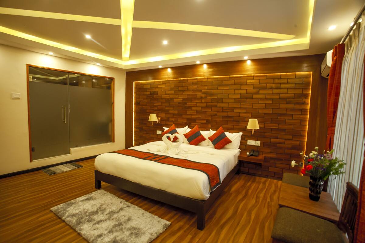 A modern bedroom with a brick-patterned headboard, a large bed with white bedding and decorative pillows, a bedside table with a lamp, and a small seating area with a chair and table.