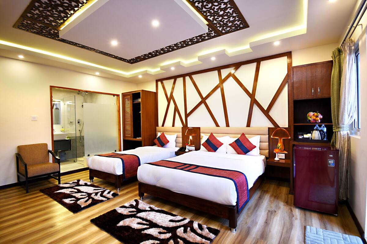 A hotel room with two beds, a sitting area, a refrigerator, and a bathroom with a glass shower enclosure.