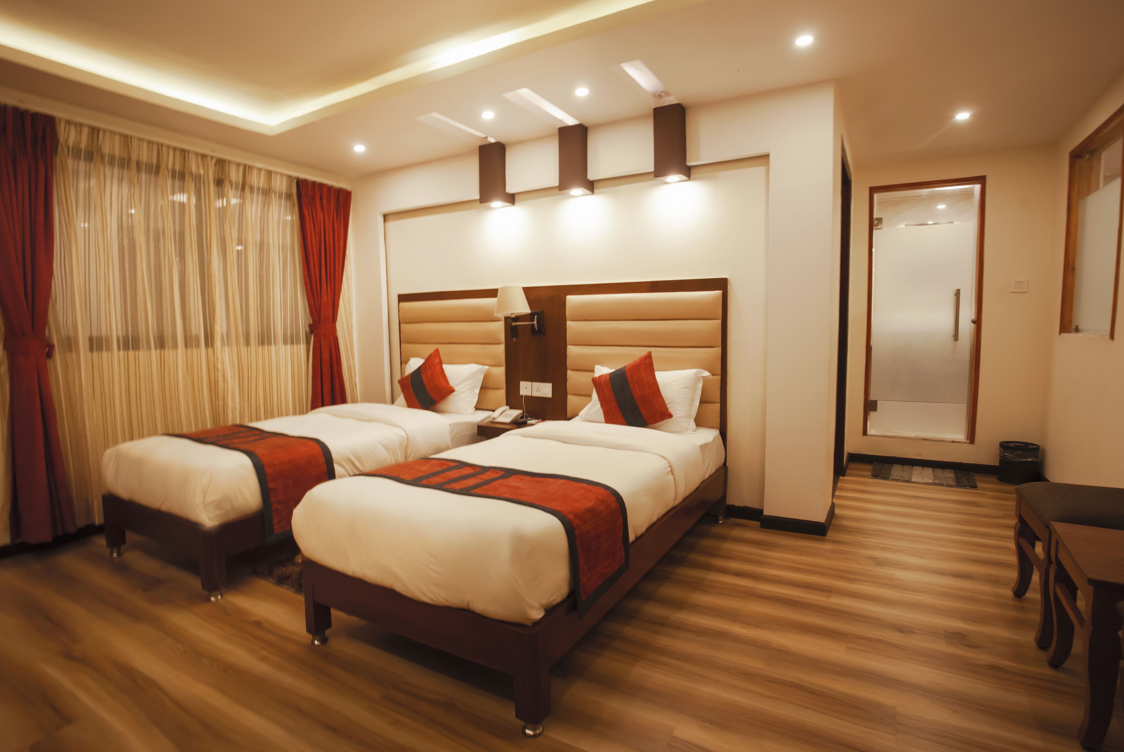 Ein Hotelzimmer mit zwei Betten, Holzboden, einem großen Fenster mit Vorhängen, einem Nachttisch mit Lampe und einer Tür mit Spiegel.