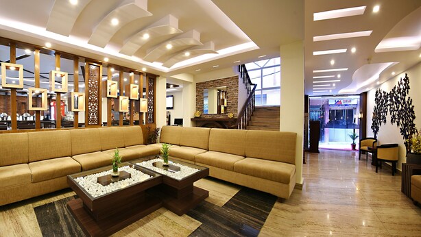 A modern lobby with a large beige sofa, two glass-top coffee tables with decorative items, a wooden partition with mirrors, and a staircase leading to another area. - Hotel Jampa (Kathmandu)