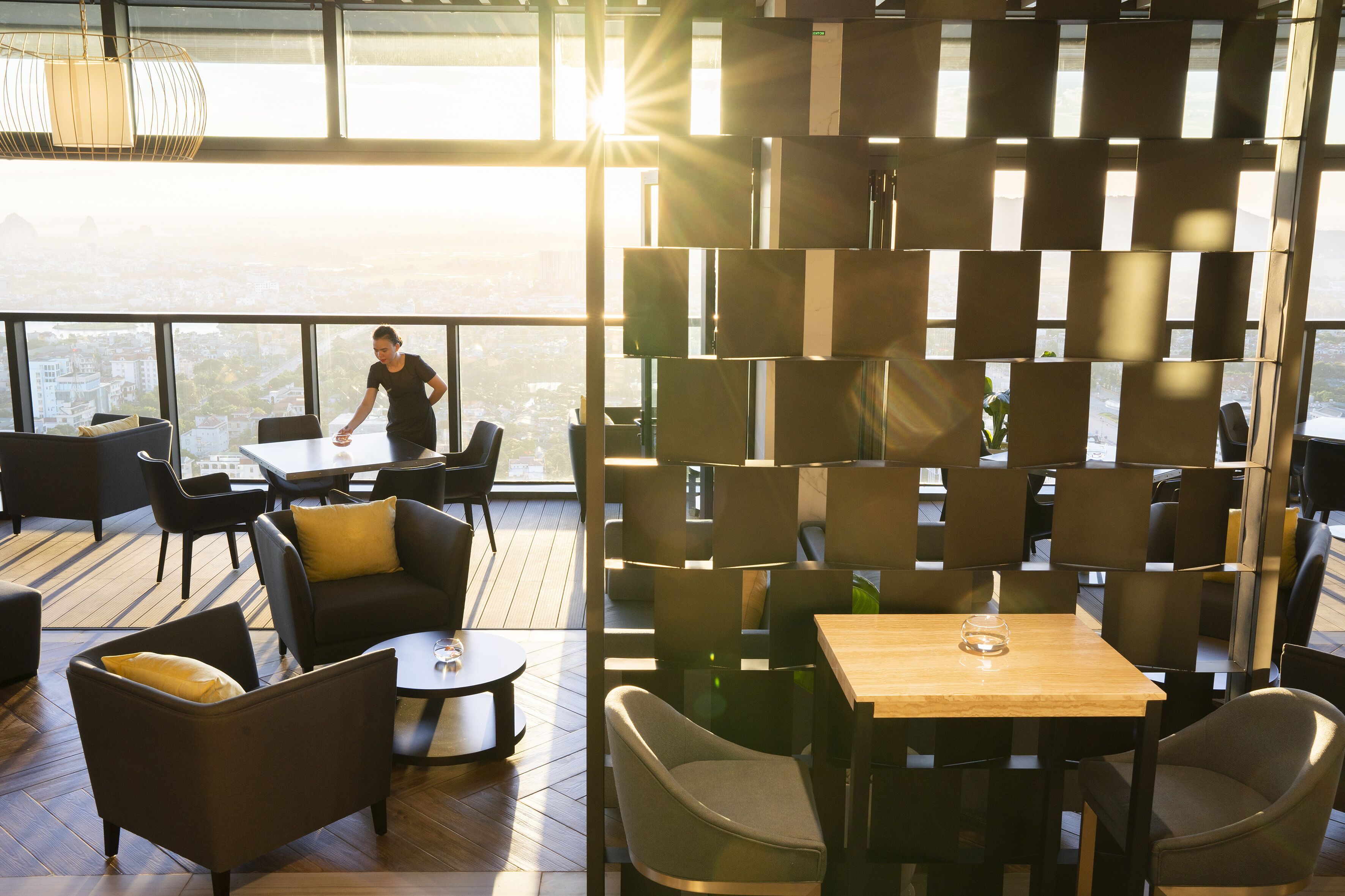 A modern interior with a high ceiling, large windows, and a view of the cityscape. There are multiple seating areas with tables and chairs.