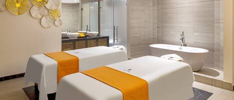 A modern spa room with two massage tables, a large mirror, a bathtub, and a decorative wall piece.