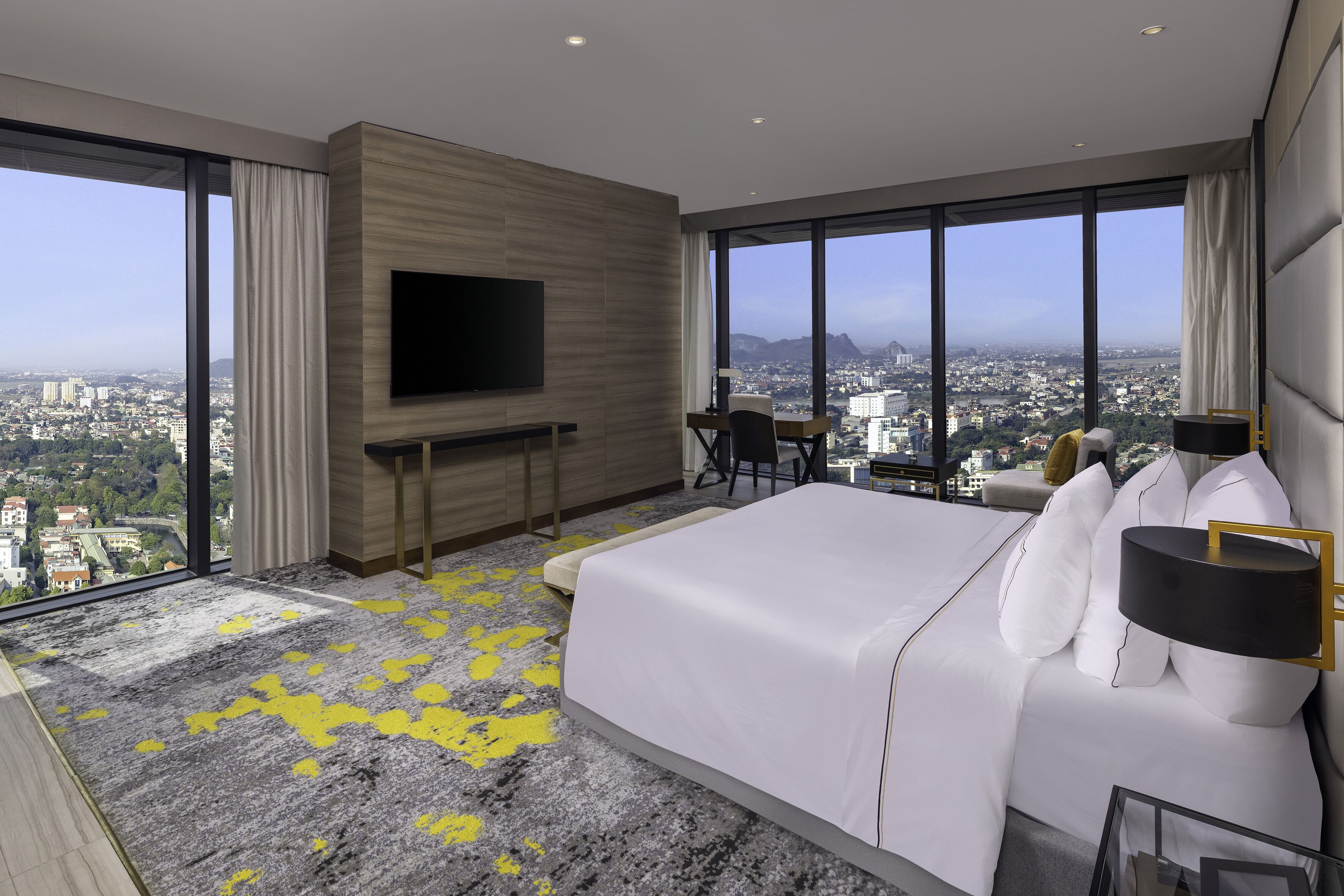 A modern hotel room with a large bed, a TV mounted on the wall, a desk, and a view of the city.