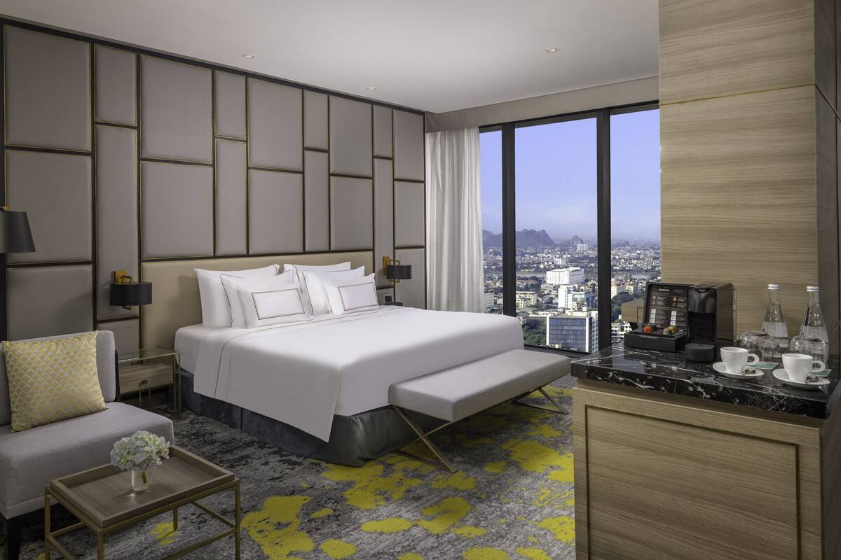 A modern hotel room with a large bed, a seating area, a small table, and a city view.