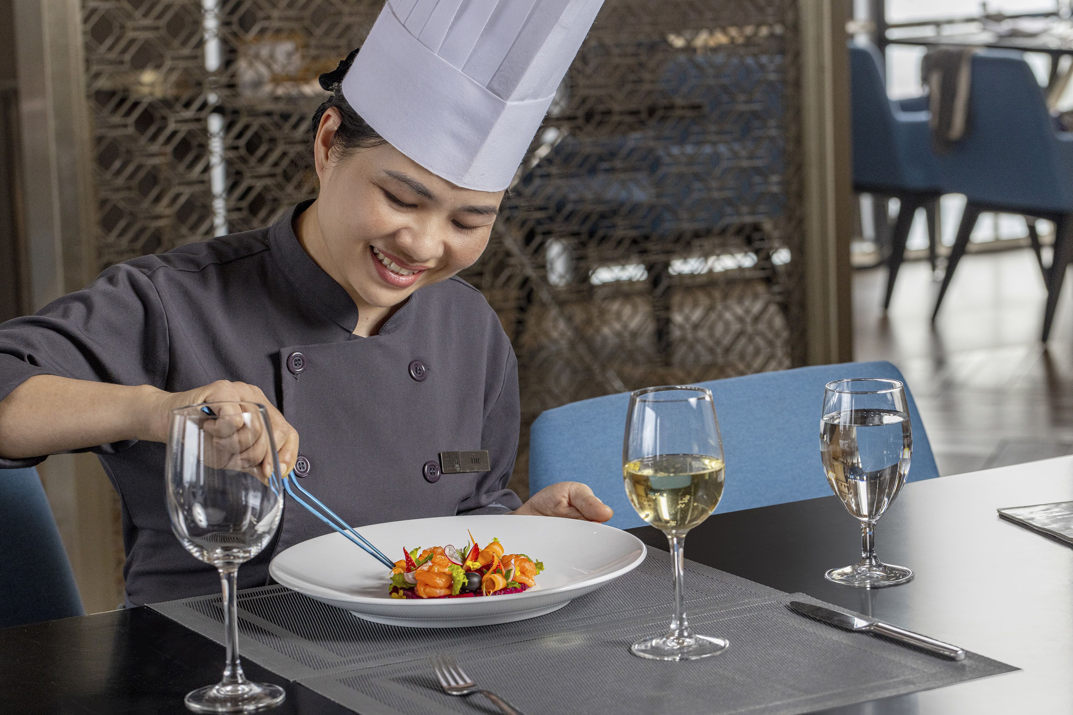 A chef in a uniform preparing a dish at a table with a wine glass and a fork.