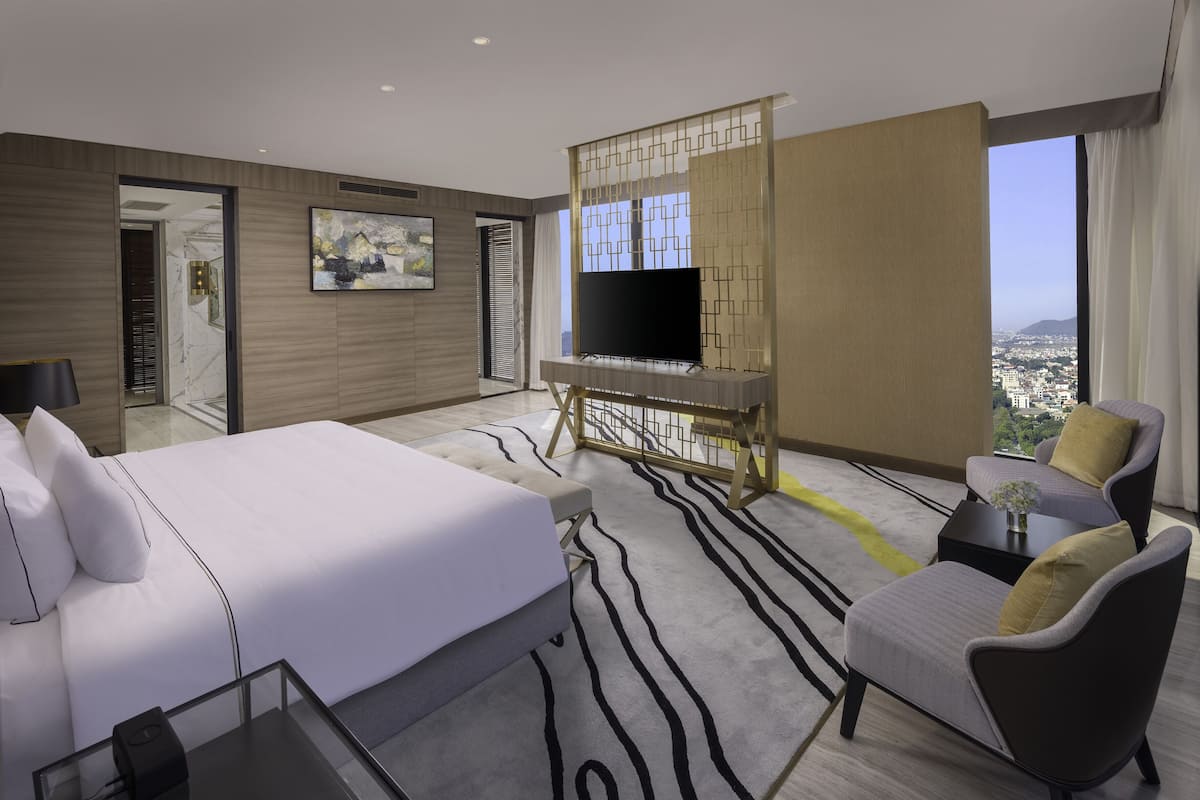 A modern hotel room with a large bed, a TV on a wooden stand, a sofa, and a view of the city.