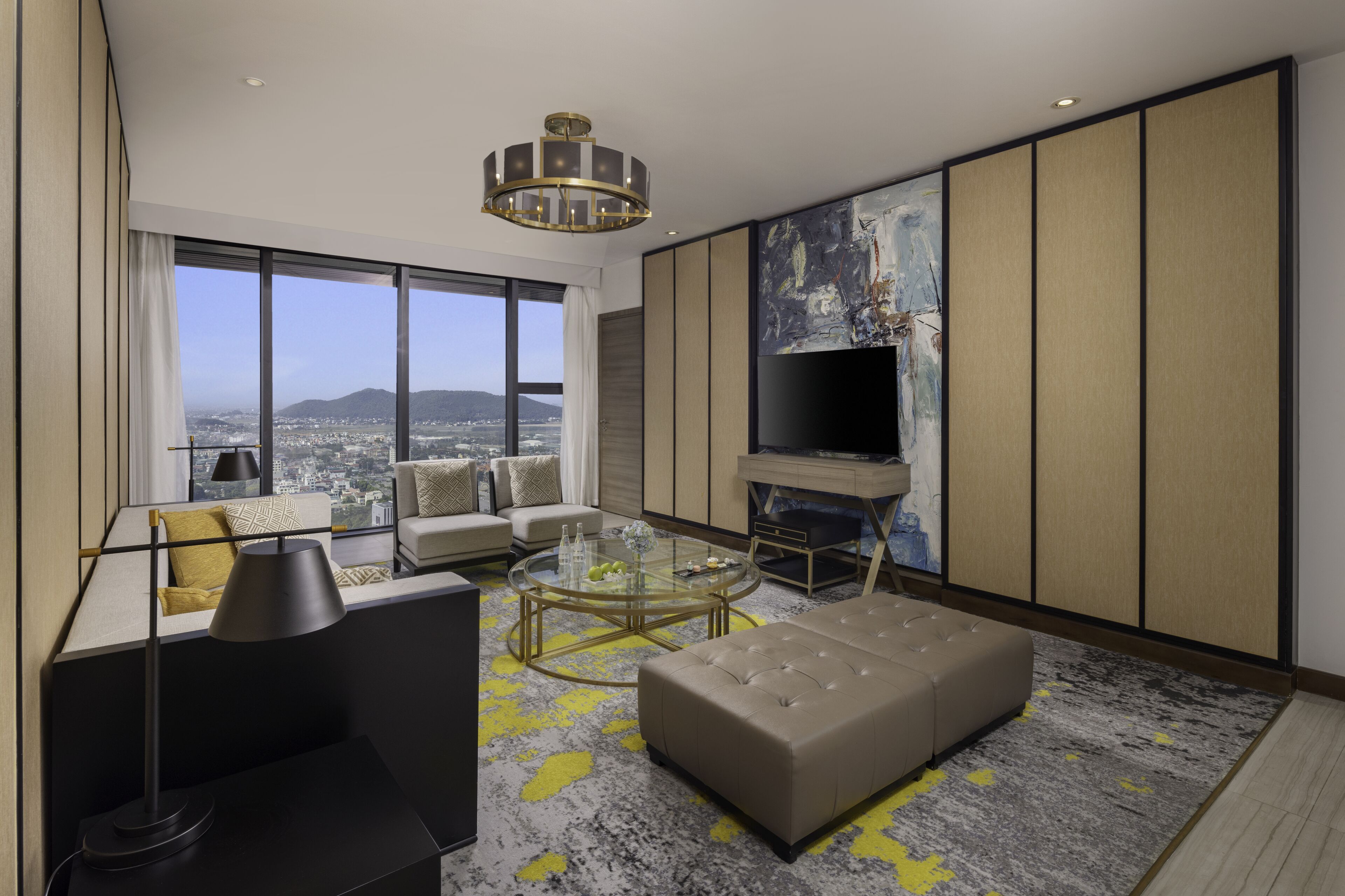 A modern living room with a large window, a round glass coffee table, a sofa, a television, and a view of the city and mountains.