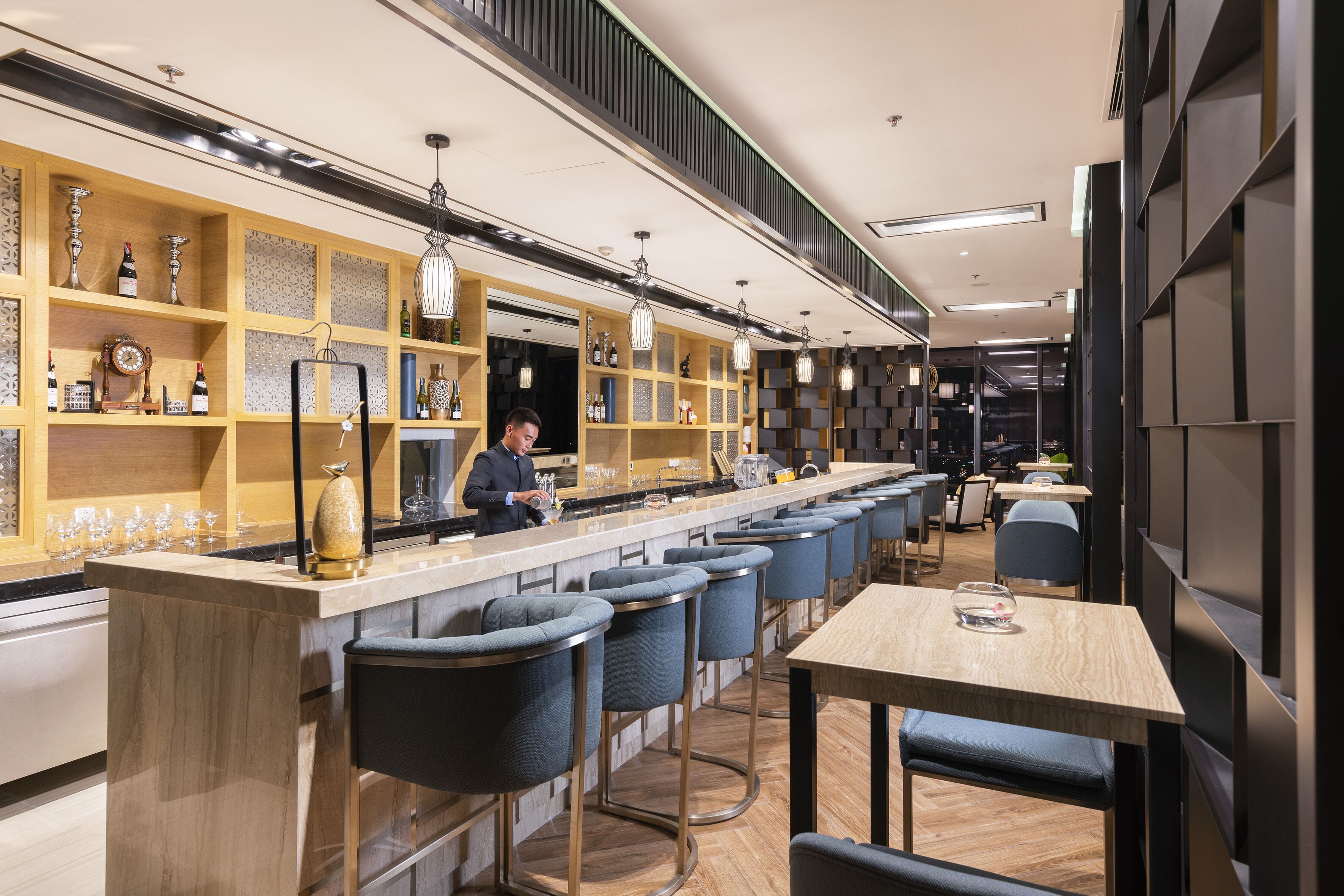 A modern bar area with a long counter, bar stools, and shelves with bottles and glasses.