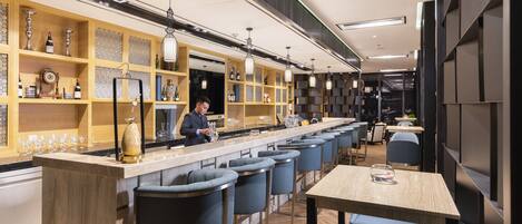 A modern bar area with a long counter, bar stools, and shelves with bottles and glasses.