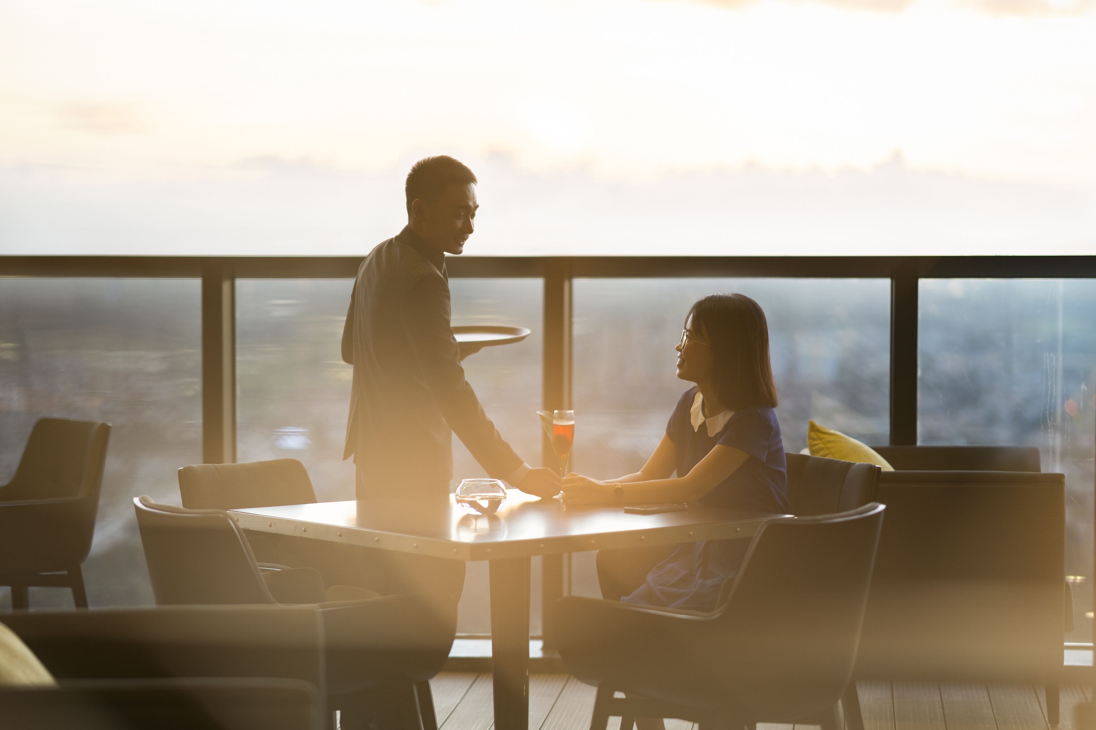 A high-rise restaurant with a panoramic city view, modern furniture, and a glass table.