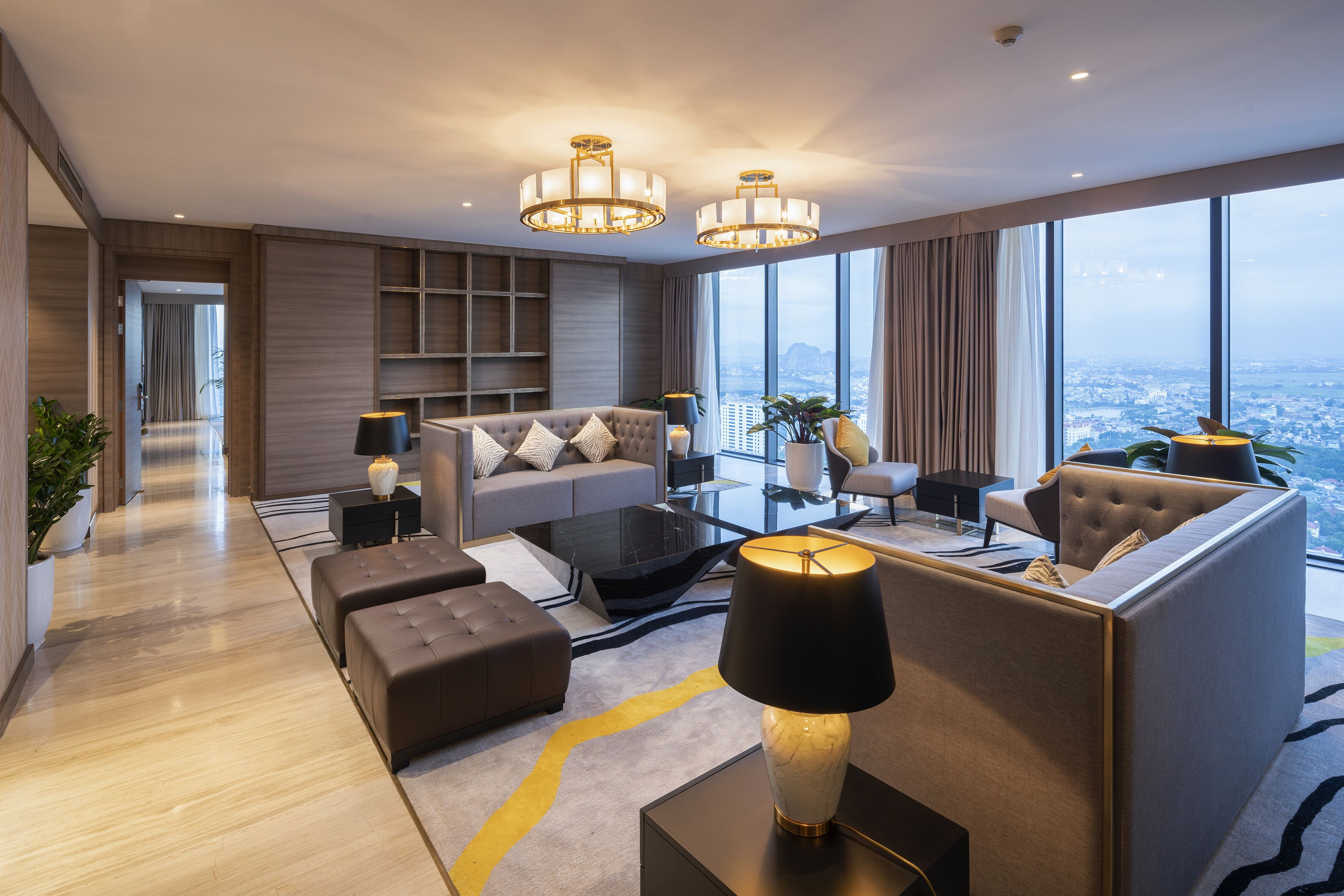 A modern hotel living room with a sofa, ottoman, coffee table, and a view of the city through large windows.