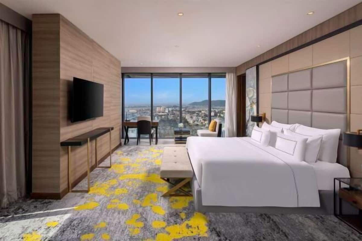 A modern hotel room with a large bed, a desk, a TV, and a city view.
