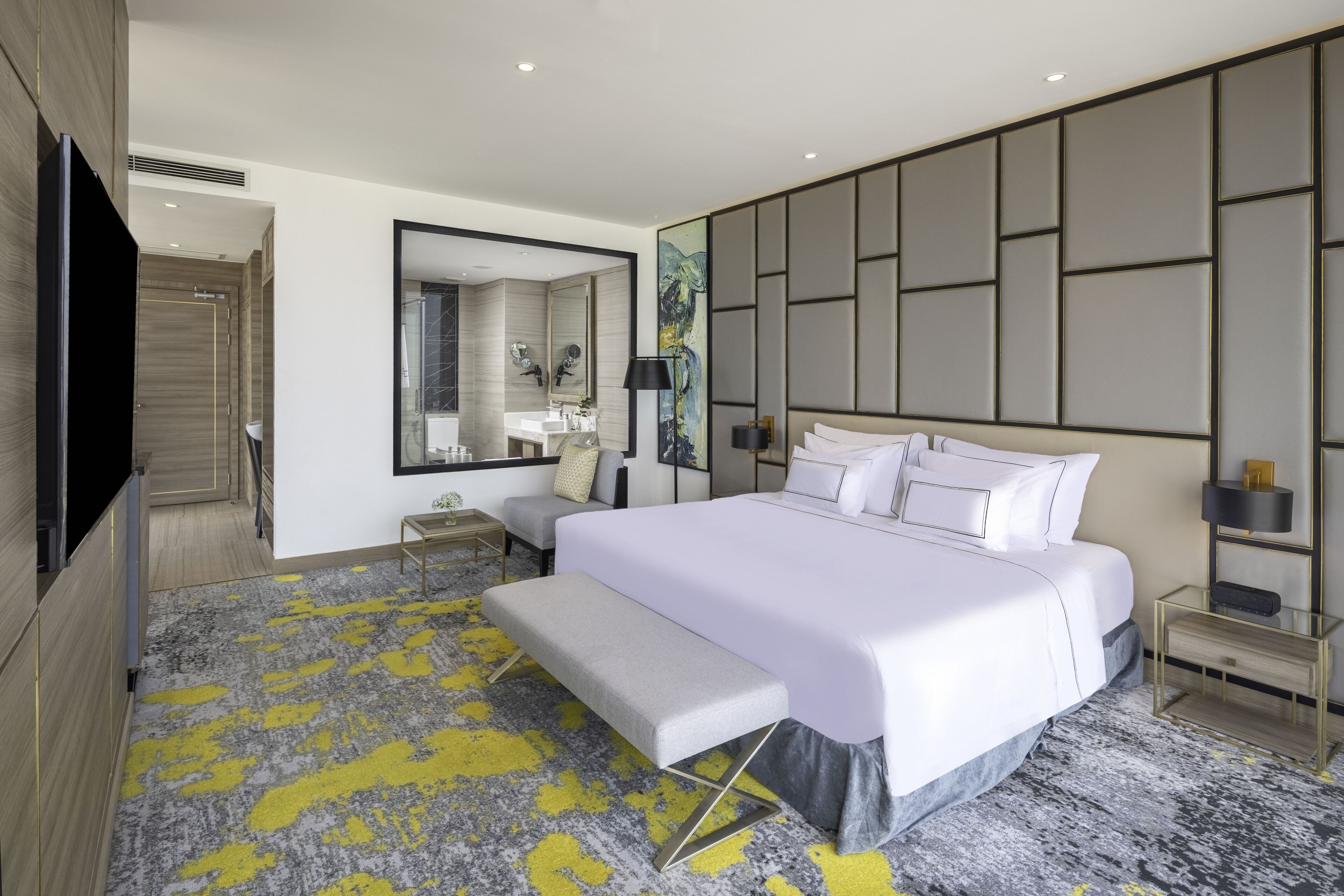 Une chambre d’hôtel moderne dotée d’un grand lit, d’une table de chevet, d’un banc, d’une télévision et d’une salle de bain avec un miroir, ainsi que d’un mur orné d’un motif géométrique.