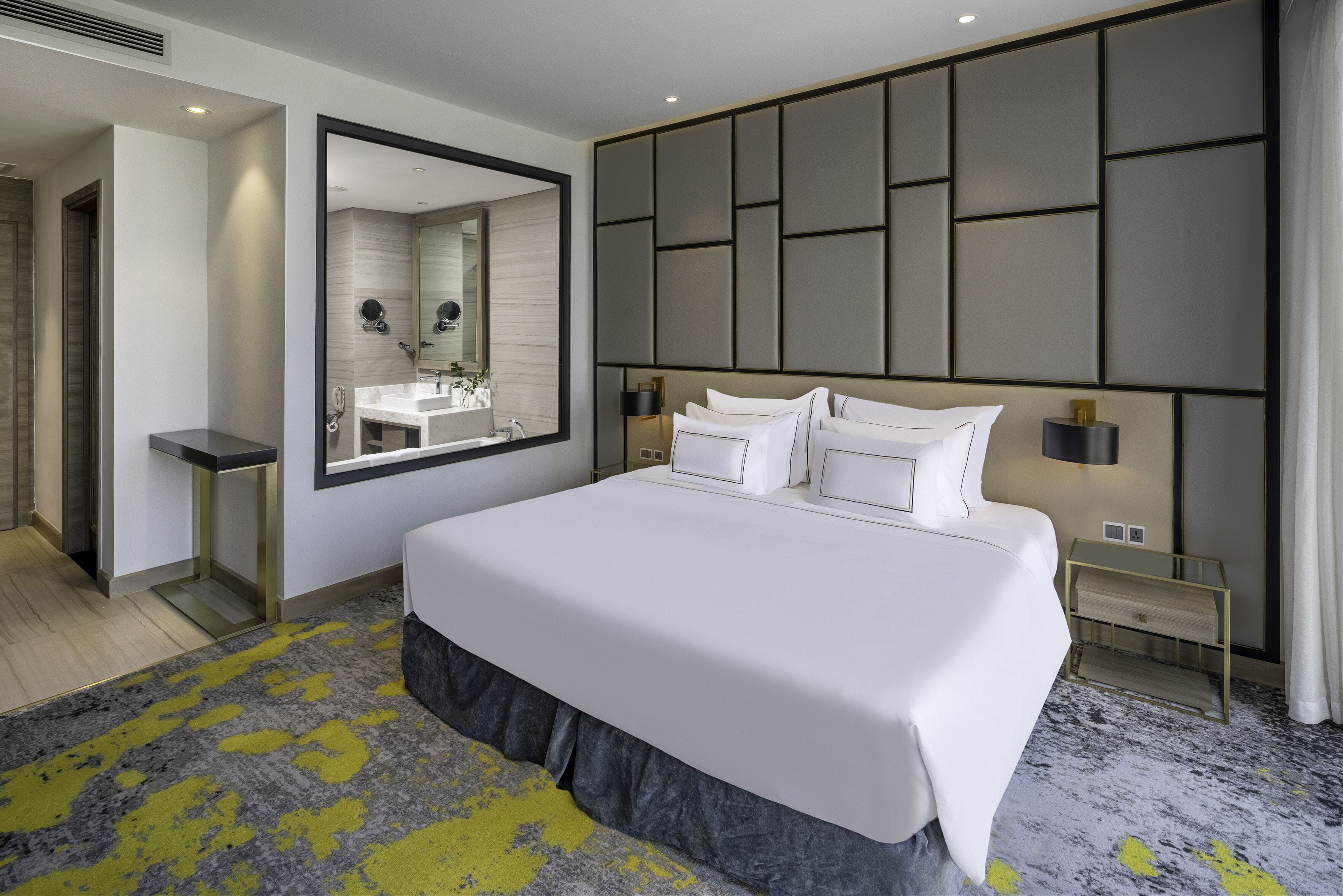 A modern hotel room with a large bed, a built-in vanity with a mirror, and a large wall panel design.