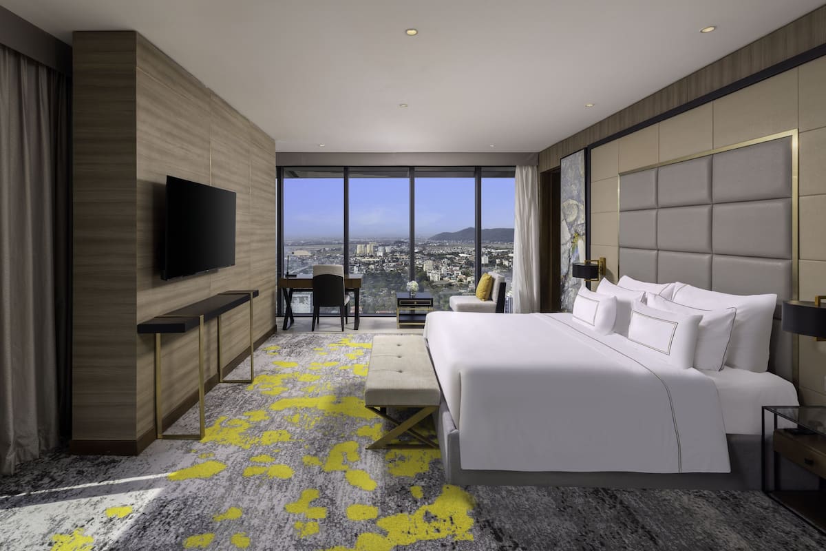 A modern hotel room with a large bed, a desk, a chair, and a TV. The room has a city view through large windows.