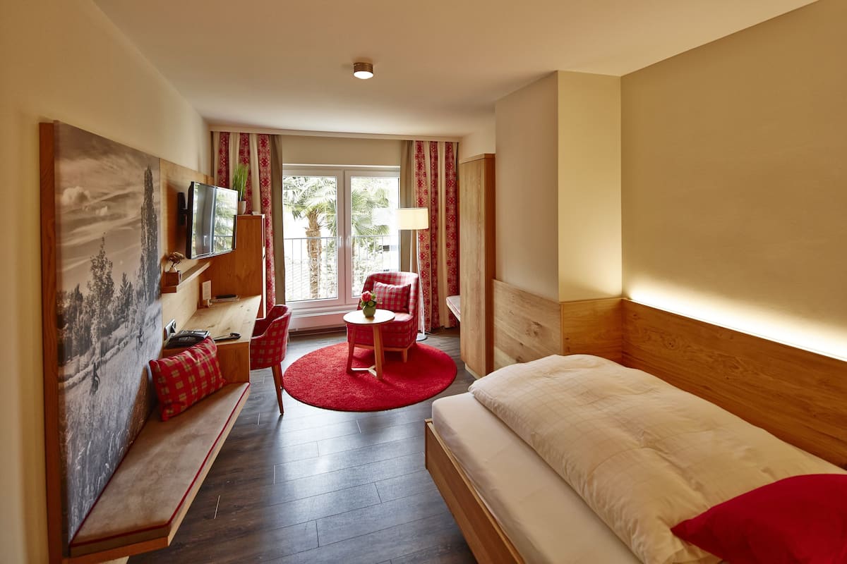 Quarto de hotel com uma cama, área de estar com um tapete vermelho, televisão e janela com cortinas.