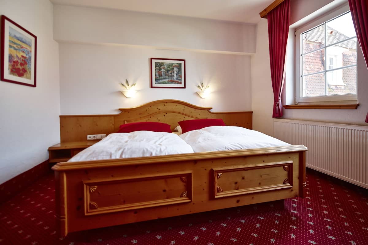 Uma cama de madeira com roupa de cama branca, duas luzes fixadas na parede e uma janela com cortinas vermelhas.