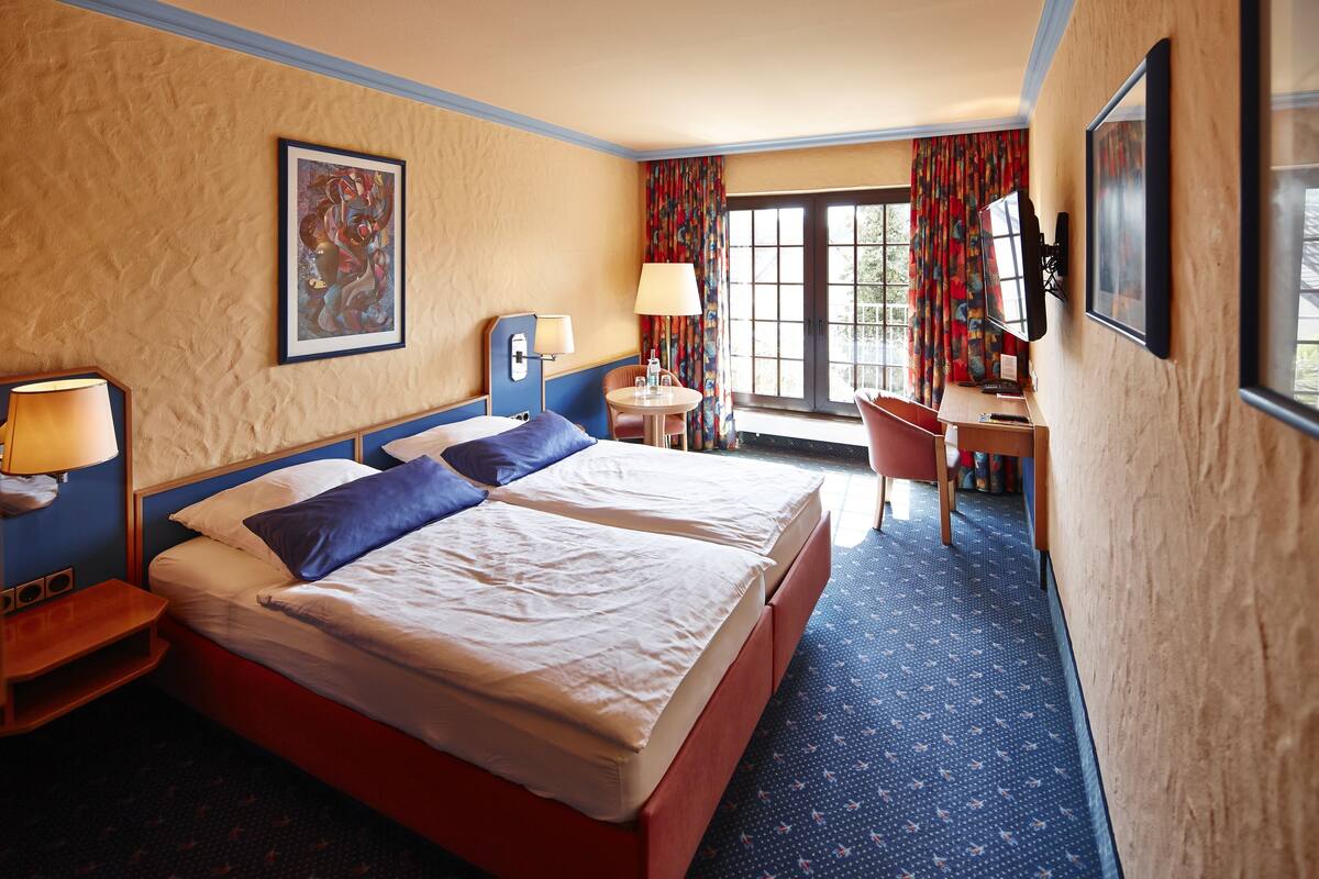 Quarto de hotel com duas camas, uma mesa, uma cadeira, uma TV e uma janela com cortinas.