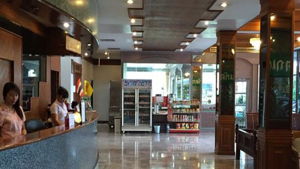 Um saguão de hotel com um balcão de recepção curvo, uma vitrine refrigerada e uma área de estar com colunas de madeira.