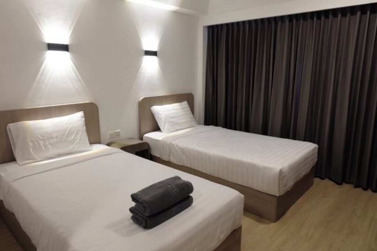A hotel room with two beds, each with white linens and a headboard. There is a folded towel on the bed in the foreground. The room has a large window with dark curtains, and the walls are painted white.